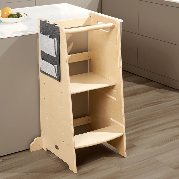 A wooden learning tower with two shelves and a bag hanging on it stands in a kitchen. Sunlight streams through the window, illuminating the space