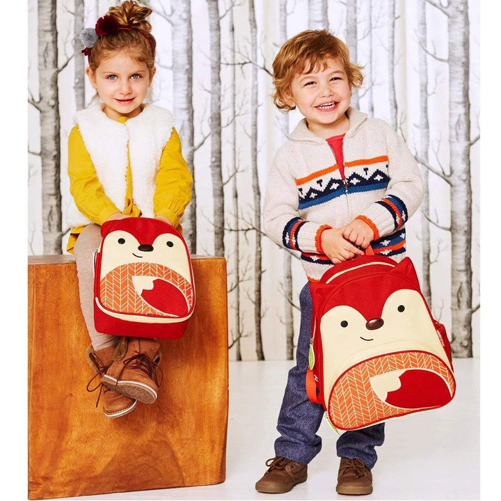 2 toddlers happily wearing a Skip Hop Zoo Little Kid Backpack in the Ferguson Fox design, walking in a park.