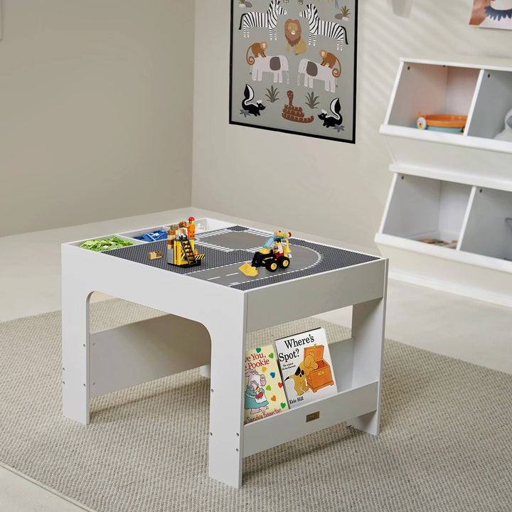 The playroom features a wooden activity table. The table features a natural finish, built-in legs, and a spacious tabletop with a road pattern made from four City Baseplate Street pieces. The table has side storage compartments and a chalkboard surface. Books are stored neatly on a shelf under the table.