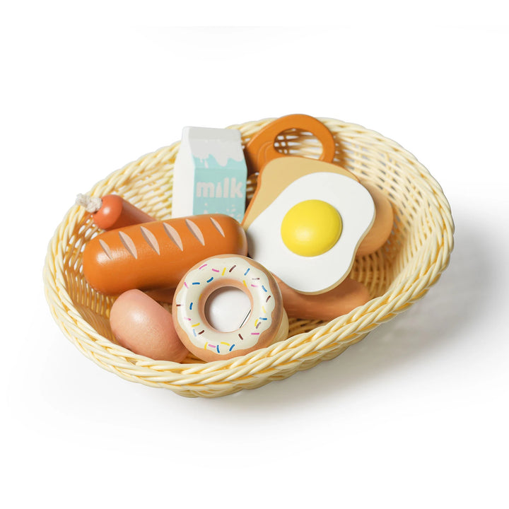 A wicker basket overflows with wooden pretend breakfast play food, including a carton of milk, eggs, bread, sausage, and a sprinkled donut.