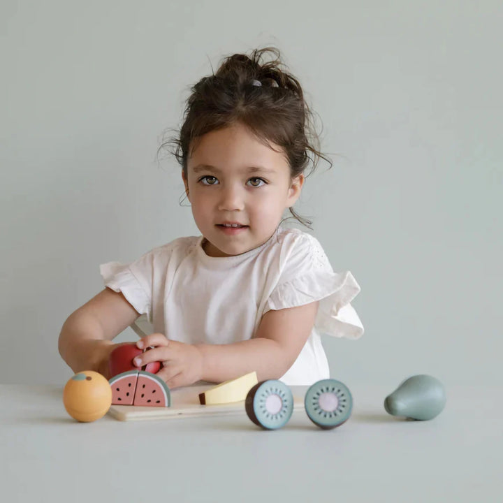 A smiling child is playing with a wooden fruit set, pretending to cut and share the different fruits with their friends or stuffed animals.